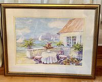 Frontal view of the watercolor painting showing the full framed artwork with visible sailboats on the water, a table, chairs, and plants in front of a stucco cottage with a thatched roof.