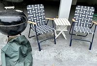 Full view showing the black Weber kettle grill with white handles, two blue folding chairs with patterned webbing, small white folding side table, and green grill cover on the ground.