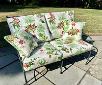 Front view of deep green aluminum garden settee with floral cushions and two throw pillows outdoors on stone patio.