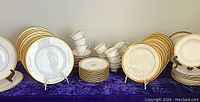 Full view of dinnerware assortment showcasing stacked plates, cups, saucers, and bowls with golden rims on dark blue cloth