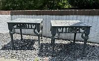 Pair of square outdoor end tables with cast iron appearance placed on gravel outside in front of white brick wall. Shows overall shape, size and condition.