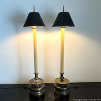 Pair of tall accent table lamps lit, showing warm golden glow from gold foil lined black paper shades, cream resin-sleeved columns, and heavy brass bases topped with pointed finials.