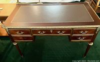 Front view of desk showing leather inset writing surface with gilt trim and dual side drawers with stylized ribbon metal pulls, one pull missing.