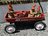 Red ACE Hardware Radio Flyer metal wagon on gravel with three garden gnomes and one toy tractor inside, viewed from above front angle.
