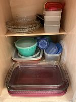 Wide view of shelf with stacked glass pie plates, bowls with lids, plastic food storage containers, and two glass baking pans on red mats