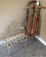 Photo shows child sized white metal bench with ornate scrollwork and a wooden Royal Racer sled propped against the wall.