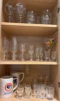 Full view of three shelves inside a wooden cabinet showing an assortment of glassware including beer mugs, stemmed glasses, shot glasses, and ceramic mugs.