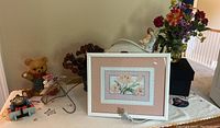 Wide shot of bedroom decor items on table including framed floral print, teddy bear figurine, faux flowers, and toy train