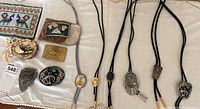 Overview photo showing multiple bolo ties with braided cords, and belt buckles on a fabric surface.