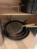 Stack of baking pans and cookware including red saucepan, black pan with wooden handle, nested pans, and black speckled roasting pan