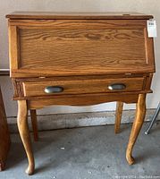 Closed view of wooden secretary desk showing wood grain finish, drawer with metal pulls, and curved cabriole legs.