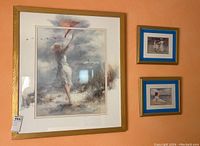 Four framed art prints displayed on peach wall, including one large with woman flying kite and three smaller beach scenes plus a white hat.