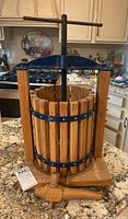 Front view of wooden fruit press with vertical slats, metal bands, wooden base, top screw handle, and front spigot for juice.