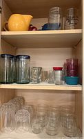 Wide view of cabinet shelf with assorted glassware showing yellow teapot, clear mugs and jars, small cups, and red/purple tumblers.