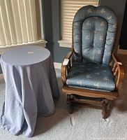 Wooden glider chair with button tufted blue floral upholstery and round side table covered in gray linen cloth next to it.
