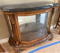 Front angle view of wooden credenza showing the green stone top, curved glass front panel, and ornate carved wood base and legs.