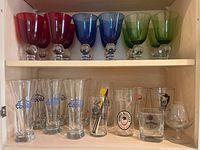 Top shelf with colored stemmed drinking glasses red, blue, green with clear rounded bases and bottom shelf with assorted pint and drinking glasses including Coors and other beer brand logos.