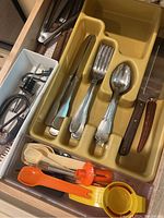Drawer with silver-tone flatware including knives, forks, spoons, and orange and yellow plastic peelers, a wine bottle opener and other small tools.