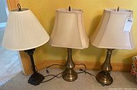 Three table lamps positioned side by side on carpet against a yellow wall; two lamps have matching brass bases and beige rectangular shades, third has a dark base with a white pleated shade.
