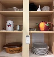Front view of two kitchen cabinets showing varied kitchenware including vintage Pyrex amber baking dish with brown lid, ceramic apple teapot, cherry jar, round containers, stacked plates, plastic containers, funnel, and black lid.