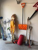 Garage corner showing orange shovel, brown spade, red rake, smaller spade, red rolled item, scarecrow, broom, metal sign, and red leaf blower mounted on wall.