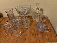 Wide shot showing all pressed glass items on wooden table, including clear and lavender glass pieces such as goblet, creamers, compotes, decanter and lidded container.