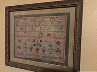 Framed embroidery sampler displayed full view showing alphabet letters, numbers, and decorative floral and geometric designs with signature and date at bottom.