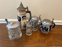 Five antique German beer steins displayed on wooden surface showing various sizes, materials, and decorated pewter lids.