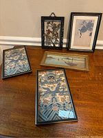 Overview photo showing all framed artworks on a wooden surface including the embroidery and prints.