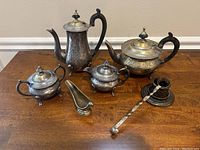 Photo showing engraved brass coffee pot, teapot, two sugar bowls with lids and handles, creamer, gravy boat, and a ladle with twisted handle and holder, all with patina and aged look.