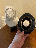 The bust on a wooden surface next to a hand holding the black oval frame with a cameo inside.