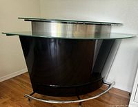 Front view of bar showing black curved body, thick clear glass top, metal frame footrail