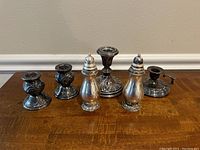 Photo displaying four weighted silver plate candlesticks and one pair of silver plate salt and pepper shakers arranged on a wooden surface.