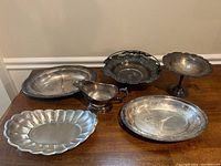 Six silver-plated serving pieces on wooden table showing trays, gravy boat, and compote bowls with tarnish and wear.