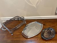 Three vintage silver-plated serving trays: rectangular glass-top tray with ornate silver sides and claw feet, oval tray with wooden handles, and small round tray with scalloped edges.