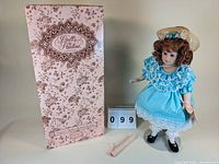 Full view of the porcelain doll standing next to her original box, showing the doll with curly auburn hair, blue dress and hat.