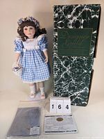 Photo showing the doll dressed in a blue and white gingham dress with white apron and floral headband, standing next to original green marbled box, certificate, and paperwork.