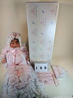 Photo showing the Lee Middleton doll dressed in the pink lace gown and bonnet, original box, certificate of authenticity, and number card 169.