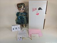 Photo of doll in plastic wrap standing upright next to original white box with Adora Belle logo and face illustration.