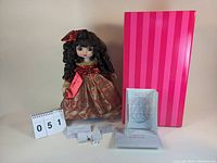 Photo showing the Marie Osmond Adora Belle Holiday 2004 doll dressed in a red and gold plaid holiday dress with lace, curly dark hair with red bow. The doll stands beside a large pink striped box and several papers, including the certificate of authenticity.