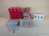 Photo showing the doll seated with bubble wrap on legs, original pink striped box behind, and certificate of authenticity with necklace displayed in front.