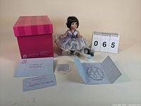 Photo of the doll seated next to its original pink and purple striped box, certificate of authenticity booklet, small potted plant, and lot number card.
