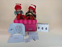 Two Marie Osmond collectible dolls named Jingles and Belle seated on a pink box with accompanying certificates and tags laid out.