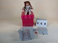 Full view of the doll seated on its original pink and red striped box, with certificate and small accessories displayed