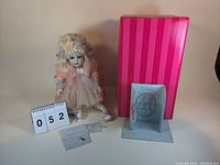 Photo of the seated Arabella doll with pink tulle dress, floral headpiece, and matching shoes, alongside the pink striped box and the certificate of authenticity displayed upright.