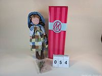 Photo shows the full doll standing next to its box, a small bag of accessories is placed in front and a number card '054'