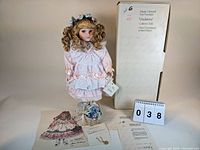 Photo shows the doll standing with long curly blonde hair, dressed in a pink and white dress, accompanied by original box, doll stand, certification and bracelet.