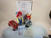 Photo of two Marie Osmond limited edition 'Miracle Children' dolls standing with a brown plush Knickerbocker bear sitting in a red wooden wagon, accompanied by documents and a numbered card 034.