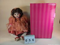 Photo showing the doll in peach dress seated next to the original pink striped box and lot number display.