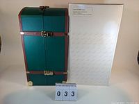 Closed green trunk with brown leather accents, silver clasps next to a white box with Marie Osmond print.